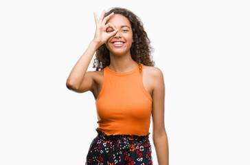 Young hispanic woman with happy face smiling doing ok sign with hand on eye looking through fingers