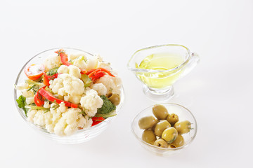 Fresh salad of cauliflower with tomato, vegetables, broccoli, greens and olives in a glass bowl with a glass nipple on a white background.