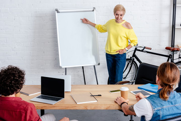 smiling young woman pointing at blank whiteboard and looking at male colleagues in office