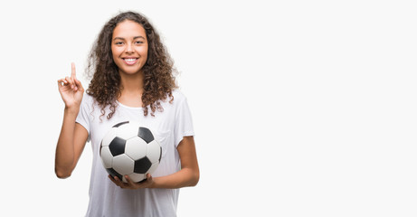 Young hispanic woman holding soccer football ball surprised with an idea or question pointing finger with happy face, number one