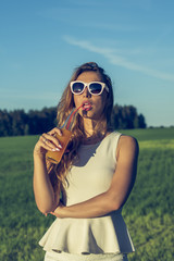 A young slender girl with glasses with curly hair smiles and drinks an alcoholic or non-alcoholic cocktail through a straw from a bottle on a sunny summer day with sexy lips