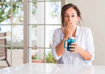 Middle aged woman drinking milk shake in a glass cover mouth with hand shocked with shame for mistake, expression of fear, scared in silence, secret concept