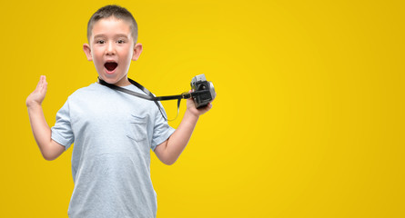Dark haired little child holding vintage camera very happy and excited, winner expression celebrating victory screaming with big smile and raised hands