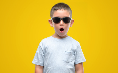 Dark haired little child wearing sunglasses scared in shock with a surprise face, afraid and...
