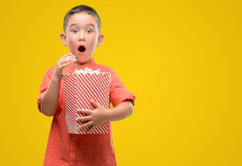 Dark haired little child eating popcorn scared in shock with a surprise face, afraid and excited with fear expression
