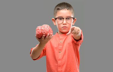 Dark haired little child with glasses holding a brain pointing with finger to the camera and to...