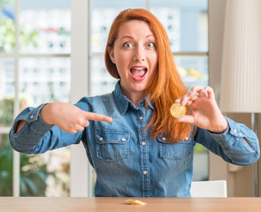 Redhead woman holding bitcoin cryptocurrency at home very happy pointing with hand and finger