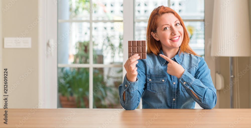 Wall mural Redhead woman holding chocolate bar at home very happy pointing with hand and finger to the side