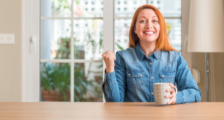 Fototapeta premium Redhead woman holding a cup of coffee screaming proud and celebrating victory and success very excited, cheering emotion
