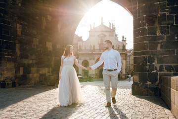 Sensual romantic couple of groom and bride in beautiful wedding dress walking holding hands under the stone arch on sunset. Under the Charles bridge. Prague Czhech republic.