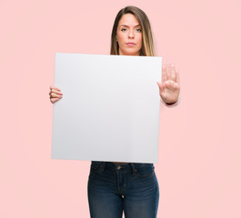 Fototapeta na wymiar Beautiful young woman holding advertising banner with open hand doing stop sign with serious and confident expression, defense gesture