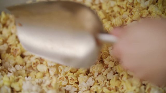 Human hand pouring ready popcorn from popcorn machine by ladle. Process of popcorn production. Selling fresh pop corn in movie theatre. Machine for production of corn. Food cinema background