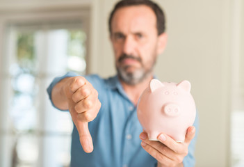 Middle age man save money on piggy bank with angry face, negative sign showing dislike with thumbs...