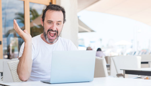 Handsome senior man using laptop at restaurant very happy and excited, winner expression celebrating victory screaming with big smile and raised hands