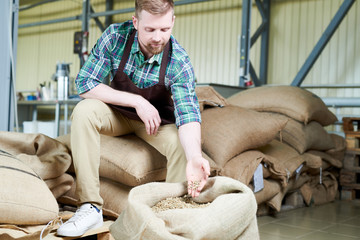 Full length portrait of handsome young man sitting on heap of  burlap bags with coffee, gently...