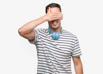 Fototapeta premium Handsome young man wearing headphones smiling and laughing with hand on face covering eyes for surprise. Blind concept.