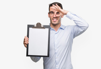 Handsome young business man showing a clipboard with happy face smiling doing ok sign with hand on eye looking through fingers