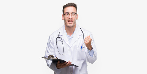 Handsome young doctor man holding a clipboard screaming proud and celebrating victory and success very excited, cheering emotion