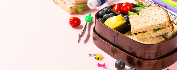 Healthy wooden lunch box with sandwiches, vegetables and fruits on pink background and school stationery. Childrens eating concept with copy space.