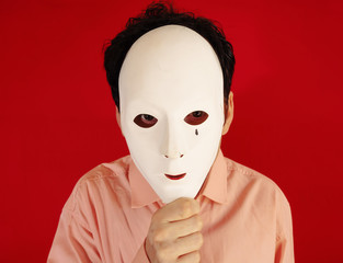 A man hiding behind an ugly plastic mask, eyes scanning the surroundings. Red background.
