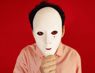 A man hiding behind an ugly plastic mask, eyes scanning the surroundings. Red background.
