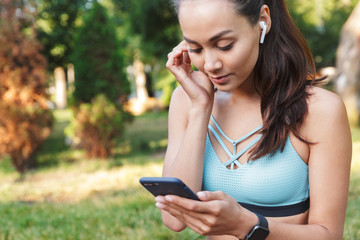 Image of beautiful caucasian woman 20s in sportswear using smartphone and bluetooth earbud, while...