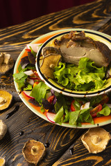 Serve a large piece of pork steak, decorated with lettuce leaves, herbs, slices of cherry tomatoes, radishes, and dried mushrooms in plates on an old wooden table.