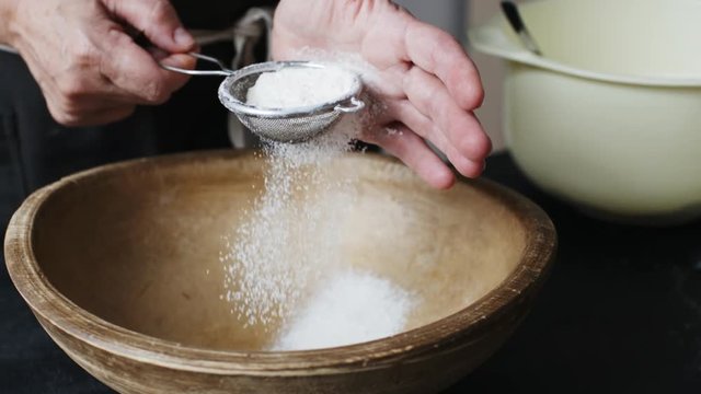 Slow motion shot of aged female hands sifting flour by sieve in wooden bowl.