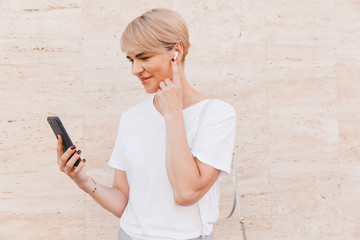 Photo of happy blond woman wearing white t-shirt using mobile phone, while standing against beige...