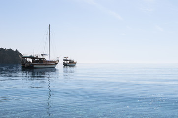 Fototapeta premium Small yacht and boat in the blue calm sea under cloudless sky in sunny morning; Sailboat with folded sails; Reflection of ships in calm water; Seascape with two boats