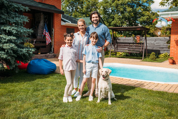 cheerful family with labrador dog looking at camera while standing on backyard of country house on summer day