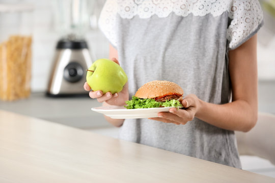 Woman holding tasty sandwich and fresh apple over table. Choice between diet and unhealthy food