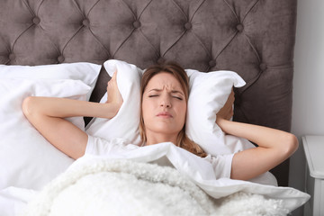 Young woman covering ears with pillow while trying to sleep in bed at home. Early morning