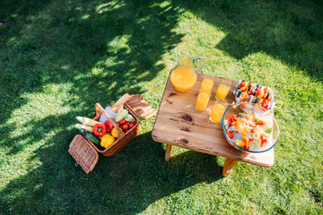 top view of basket with food for picnic, glasses of  juice and fresh salad on wooden chair on green lawn