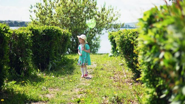 A little sweet girl in a blue dress is catching insects with a green net in the alley next to the river on a sunny summer day.