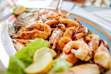 Delicious fresh seafood mix: dorado fish, shrimps, octopus and squid with lemon and salad leaves on the metallic plate. Tasty Mediterranean dish on the table. Close up view.