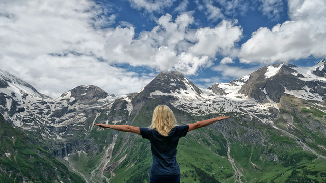blonde Frau in Rückenansicht breitet die Arme aus und blickt auf ein Hochgebirge mit blauem Himmel in den Alpen