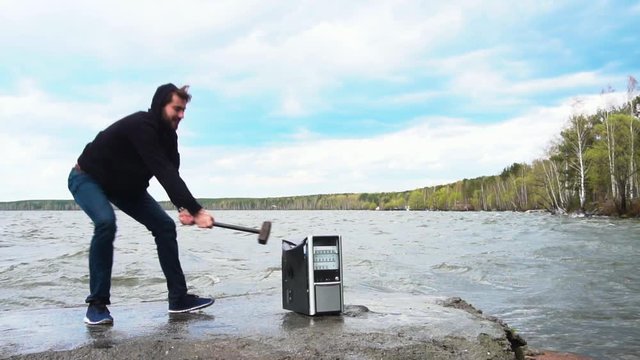 Concept of freedom from computer addiction - Man breaks system unit of a computer with a hummer outside, lake with forest background. Angry man destroying a laptop or computer with a hammer.