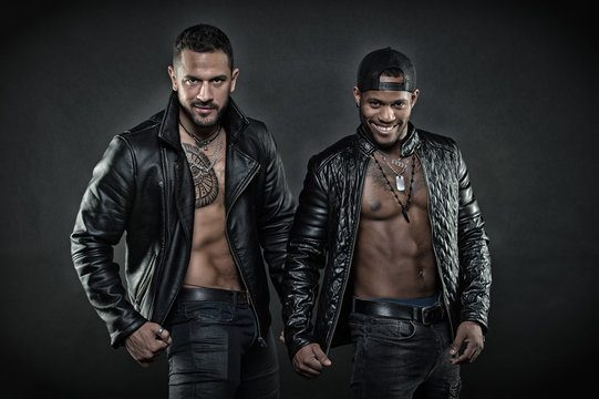 Two men in leather jackets on black background. Brutal Hispanic man with geometrical tattoo. African man with happy smile wearing cap backwards. Models with sexy muscular bodies, fitness concept