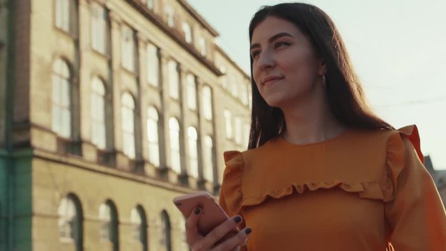 Low angle shot of young long-haired woman walking down the busy city-street, looking around and using her smartphone. Modern technologies, blogger, insta blogger. Social networks Female portrait, sun