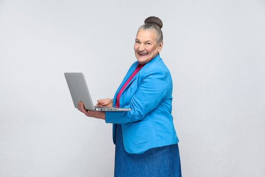 Aged woman standing and holding laptop and looking at camera with smiley face. Grandmother in light blue suit and collected gray hair bun hairstyle. indoor, Studio shot, isolated on gray background