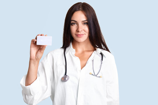 Adorable lovely female family medicine physician, wears lab coat and phonenodoscope, holds white small card with blank space for your advertising content, isolated over light blue background