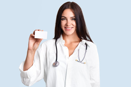 Beautiful professional female therapist wears white medical coat, stethoscope, holds blank card, smiles gently, isolated over light blue background. Medical worker models indoor. Medicine concept