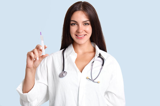 Beautiful young female doctor in white robe, has phonendoscope on neck, holds syringe with liquid, isolated over blue background, has cheerful expression. Glad woman nurse makes injections to patient