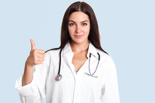 Successful talented female therapist raises thumb and shows ok sign, dressed in white robe, looks seriously at camera, stands against blue background. Medicine, people, body language concept