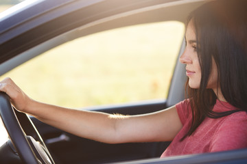 Fototapeta na wymiar Photo of attractive brunette female driver with confident expression looks at windshield, enjoys high speed and good roads, travels by car, has attractive appearance. People and transport concept