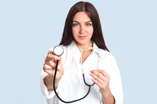 Isolated shot of prett confident female doctor advertises her new phonendoscope, ready to examine people, hear lungs and heart, isolated over blue background. Focus on stethoscope. Medical worker