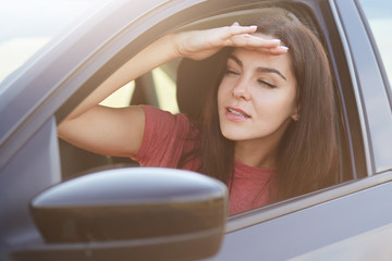 Attractive focused female keeps hand on forehead, looks into distance from car window, tries to...