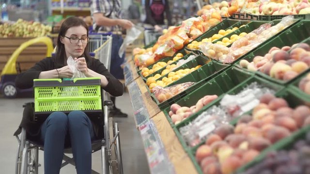 Woman with a disability in a wheelchair shopping in the supermarket chooses fruits and puts them in a package.