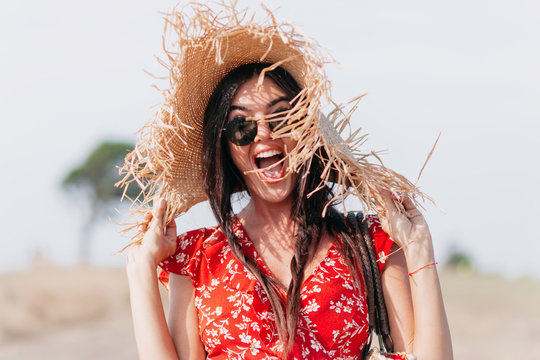 Close-up portrait of excited lovely woman wears summer dress has fun outdoors, screming with smile and covering face with hat, true happy emotions, stylish woman on vacation